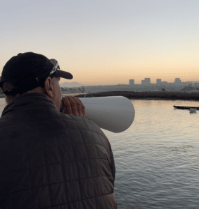 Women's Head Coach Mike Homes poses with his megaphone, facing away from the camera, in front of the Newport Harbor skyline.