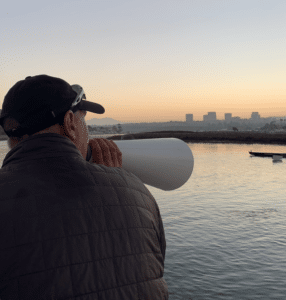 Women's Head Coach Mike Homes poses with his megaphone, facing away from the camera, in front of the Newport Harbor skyline.